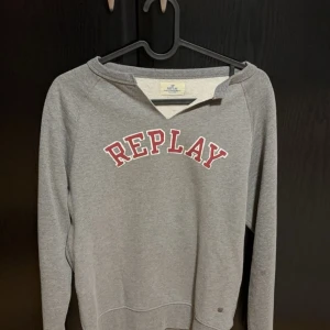 Grå tröja från Replay - Säljer en grå tröja från Replay med röd text på bröstet. Tröjan har en v-ringad hals och långa ärmar. Perfekt för en avslappnad stil.