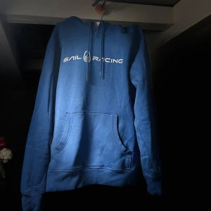 Blå hoodie från Sail Racing - Säljer en snygg blå hoodie från Sail Racing med vit logga på bröstet. Tröjan har en klassisk känguruficka och justerbar huva med dragsko. Perfekt för en avslappnad stil.