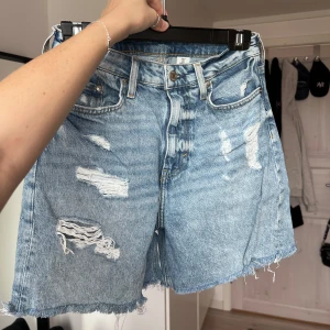 Blå jeansshorts med slitningar - Säljer ett par coola blå jeansshorts från H&M med slitna detaljer och råa kanter. Perfekta för en avslappnad stil. De har en klassisk femficksdesign och knappgylf. Storlek 36 💛 Önskar du veta exakta mått? Tveka inte att höra av dig!