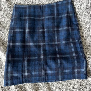 Blå rutig kjol - Snygg blå rutig kjol med en klassisk design. Kjolen har en diskret slits baktill och är perfekt för en stilren look. Den är gjord i ett mjukt material som ger en bekväm passform. Inga tecken på användning