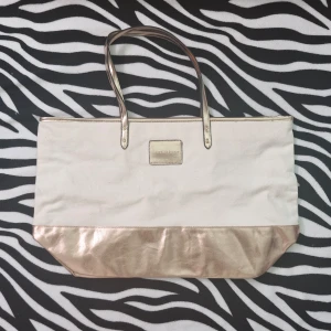 Victoria secret väska - Säljer en original vs väska. På bilden ser väskan ut lite att vara guld men egentligen är den rosegold. Väskan har knappt blivit använd men på sista bilden kan man se några, små anmärkningar på grund av att den har stått i garderoben ett tag, som har gjort att den har tappat färg. Plus har väskan små prickar på den vita delen av väskan som inte går att visa. Vet ej om den var sådär från början eller om den blev så pågrund av att den har stått i garderoben länge men annars är väskan i toppskick.🩷