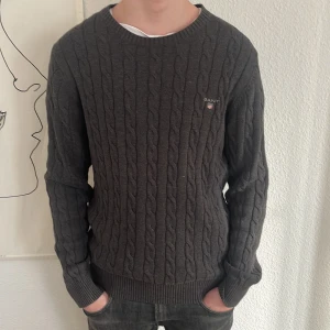 Grå kabelstickad tröja från GANT - Säljer en stilren grå kabelstickad tröja från GANT. Tröjan har en rund halsringning och långa ärmar med ribbade muddar. Passar bra till 185-190. 