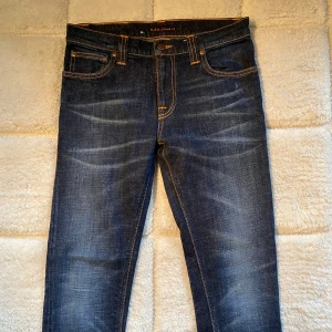 Nudie Jeans - Snygga mörkblå jeans från Nudie Jeans med orange sömmar och klassisk femficksdesign. Jeansen har en slim passform och är tillverkade i denim med en lätt tvättad look. Perfekta för en stilren vardagslook. 