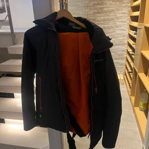 Svart jacka från 8848 Altitude - Säljer en svart jacka från 8848 Altitude med orange foder och coola märken på ärmen. Jackan har en dragkedja framtill och är perfekt för utomhusaktiviteter. Den har en sportig look med långärmade ärmar och en skön passform. Termobyxor medföljer, de är hel orange och har hängslen.