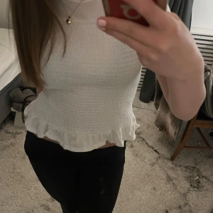 Vit topp - Säljer en söt vit smockad topp från Gina Tricot. Toppen är ärmlös och har en volangdetalj vid nederkanten som ger en feminin touch. Perfekt för varmare dagar eller som ett lager under en jacka.