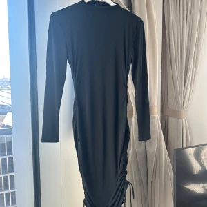 Svart klänning från Gina Tricot - Snygg svart klänning från Gina Tricot med långärmad design och åtsittande passform. Klänningen har en stilren look med rynkade detaljer på sidan som ger en extra touch. Perfekt för en chic och elegant stil.