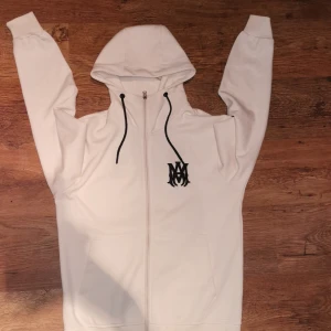 Vit hoodie från Amiri - Säljer en stilren vit hoodie från Amiri med dragkedja och svart logotyp på bröstet och ryggen. Den har en justerbar huva med svarta snören och praktiska fickor framtill. Perfekt för en avslappnad look.
