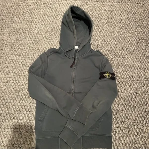 Grön hoodie från Stone Island - Säljer en snygg grå hoodie från Stone Island med dragkedja och den ikoniska loggan på ärmen. Perfekt för en stilren och avslappnad look. Hoodien har långa ärmar och en praktisk huva.