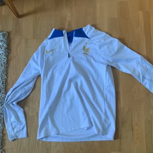 Frankrike träningskofta Nike - Snygg vit långärmad fotbollströja från Nike med blå detaljer och ett guldigt emblem på bröstet. Tröjan har en halv dragkedja framtill och är perfekt för fotbollsfans. Den har en stilren design med en modern touch.