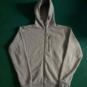 Grå hoodie från Sail Racing - Säljer en stilren grå hoodie från Sail Racing med dragkedja och en praktisk bröstficka. Perfekt för kyliga dagar med sin mjuka insida och justerbara huva. Logotypen är snyggt placerad på bröstet. Passar perfekt för en avslappnad stil.