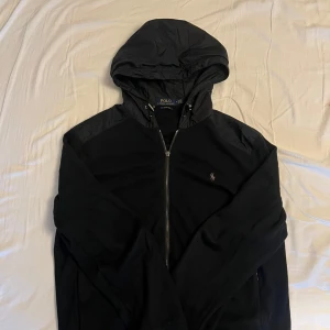 Svart jacka med huva från Polo Ralph Lauren - Snygg svart cardigan med huva från Polo Ralph Lauren. Jackan har dragkedja framtill och en liten broderad logga på bröstet. Perfekt för en stilren look.