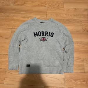 Grå tröja från Morris - Säljer en stilren grå tröja från Morris med broderad text och en Union Jack-detalj på framsidan. Tröjan har långa ärmar och ribbade muddar vid ärmslut och nederkant. Perfekt för en avslappnad och snygg look.