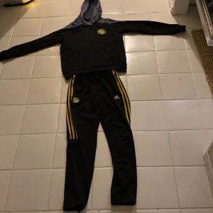 Svart och grå hoodie från Adidas - Säljer en svart och grå hoodie från Adidas med gul logga på bröstet och Stadium-tryck på ryggen. Perfekt för sportiga aktiviteter med sin bekväma passform och långa ärmar. Den har en klassisk huva och är tillverkad i ett mjukt material. Sedan ett par tränings byxor som också är från Adidas med gul logga på låret perfekt som tränings byxor när man ska iväg och träna eller på match. Perfekt för er som går i Sävehof och inte vill köpa på någon dyr hemsida 😀
