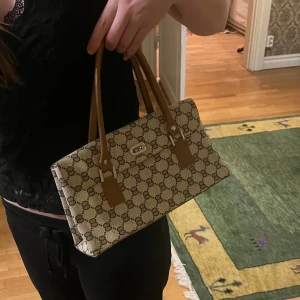 Beige och brun handväska med mönster - Snygg beige handväska med brunt mönster och bruna handtag. Väskan har en klassisk design med ett diskret märke på framsidan. Perfekt för att ge en stilren touch till din outfit.