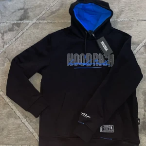 Svart hoodie från Hoodrich - Säljer en svart hoodie från Hoodrich med blå detaljer och text på bröstet. Hoodien har en blå insida i huvan och långa ärmar. Perfekt för en avslappnad stil.