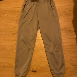 Grå mjukisbyxor från Lacoste - Snygga grå mjukisbyxor från Lacoste med elastisk midja och dragkedjefickor. Byxorna har en broderad logotyp på sidan och är perfekta för en avslappnad stil. De är tillverkade i ett mjukt material som ger en bekväm passform.