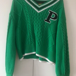 Grön stickad tröja från Gina Tricot - Säljer en grön stickad tröja från Gina Tricot med en stor svart bokstav 'P' på bröstet. Helt nytt skick aldrig använd. Tröjan har en v-ringad hals med vita ränder och långa ärmar. Perfekt för en cool och avslappnad stil. 💚