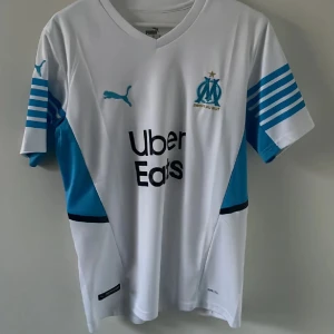 Olympique de Marseille fotbollströja Puma - Säljer en vit Olympique de Marseille matchtröja från Puma med blå detaljer och klubbmärke på bröstet. Tröjan har korta ärmar med blå ränder och sponsortryck på framsidan. Perfekt för fotbollsfans!