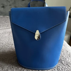 Blå axelväska från Calvin Klein - Säljer en stilren blå axelväska från Calvin Klein med vit kontrastsöm och silverfärgat lås framtill. Väskan har en unik form, justerbara axelremmar och är tillverkad i slätt skinn. Perfekt för dig som vill ha en snygg accessoar med plats för det viktigaste.. Finns defekt på baksidan se bild. Köpt för 1500kr. Kan diskutera pris vid snabb affär. Köpt 2021 och har orginal påse.