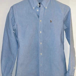 Ljusblå skjorta från Ralph Lauren - Klassisk ljusblå skjorta från Ralph Lauren med broderad logga på bröstet. Skjortan har knappar hela vägen och button-down krage. Perfekt till jeans eller chinos för en clean look.