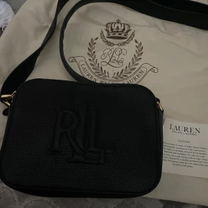 Svart axelväska från Lauren Ralph Lauren - Säljer en svart axelväska från Lauren Ralph Lauren med präglad RL-logga på framsidan. Väskan har två stora fack med guldfärgade dragkedjor och justerbar axelrem. Perfekt för att bära det viktigaste med stil.