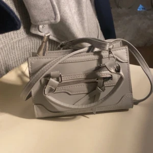 Grå axelväska med dragkedjedetalj - Säljer en stilren grå axelväska med både handtag och axelrem. Väskan har en dekorativ dragkedja framtill och tydliga sömmar som ger en modern look. Perfekt för dig som vill ha en snygg accessoar till din outfit.