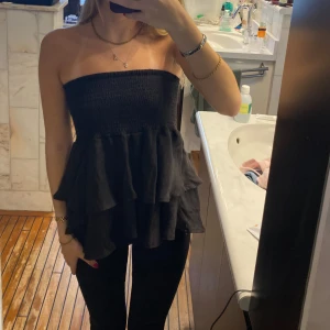 Svart volang tubtopp - Säljer en svart tubtopp med volanger. Toppen är axelbandslös och har en smockad överdel som sitter åt över bysten. Perfekt för dig som vill ha en stilren och trendig look. Funkar även som kjol 