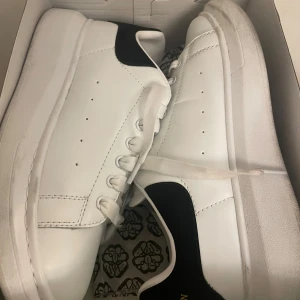 Alexander McQueen vita sneakers med svart häl - Säljer ett par klassiska vita sneakers från Alexander McQueen med svart mockadetalj på hälen och guldfärgad logga. Skorna har rund tå, platt sula och vita snören. Perfekta för dig som gillar stilrena och trendiga sneakers.