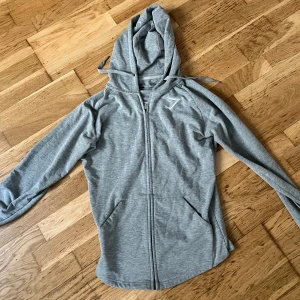 Ljusgrå hoodie med dragkedja från Gymshark - Säljer en ljusgrå hoodie från Gymshark med dragkedja framtill och huva. Jackan har långa ärmar, två fickor och en diskret Gymshark-logga på bröstet. Perfekt för träning eller chill dagar.