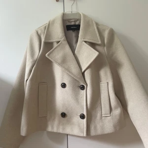 Beige kappa från Vero Moda - Snygg beige kappa från Vero Moda i storlek S med dubbelknäppning. Jackan har raka fickor framtill och en klassisk, stilren design som passar perfekt till vår och höst.