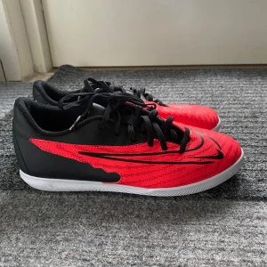 Nike Phantom GX inomhusfotbollsskor i rött och svart - Säljer ett par Nike Phantom GX inomhusfotbollsskor i rött och svart med vita sulor. Skorna har snörning och en låg profil, perfekt för inomhusspel. Snygg design med svarta detaljer och Nike-logga på sidan. I storlek 38,5. För ENDAST 250kr så får du de bästa inomhusfotbollsskorna, pris kan diskuteras.