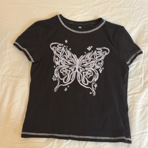 Svart t-shirt med fjärilstryck från H&M - Svart t-shirt från H&M med lila kontrastsöm och stort lila fjärilstryck på bröstet. T-shirten har korta ärmar och rund halsringning. Perfekt för dig som gillar en cool och trendig stil.❤️