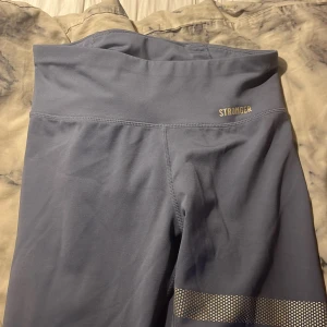 Grå träningstights från Stronger - Snygga grå -blå  träningstights från Stronger med hög midja och diskreta reflexdetaljer på benen. Tillverkade i ett stretchigt material som består av polyester och elastan. Perfekta för gymmet eller löprundan.