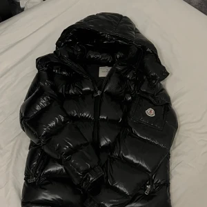 Moncler jacka - Säljer en svart, glansig dunjacka från Moncler med huva och dragkedja. Jackan har Moncler-logga på ärmen och flera fickor med dragkedja. Perfekt för kalla dagar och riktigt snygg streetstil. Defekt är ett litet hål och använd som satan därav det låga priset