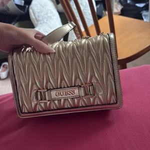 Guldig axelväska från Guess - Säljer en snygg guldig axelväska från Guess med quiltad design och kedjerem. Väskan har ett metallspänne framtill med Guess-logga och ett huvudfack med innerficka. Perfekt för att lyfta din outfit med lite extra bling!
