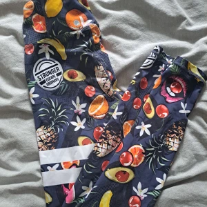 Färgglada träningstights från Stronger - Säljer ett par färgglada träningstights från Stronger med tropiskt fruktmönster i olika färger. Tightsen har hög midja och vita ränder på ena benet. Perfekta för gymmet eller löprundan.