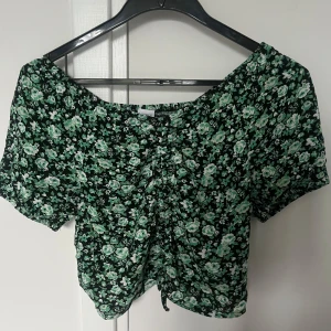 Grön blommig croppad topp från Lindex - Säljer en söt croppad topp från Lindex med grönt och vitt blommönster på svart botten. Toppen har korta ärmar och en lätt rynkad detalj framtill. Perfekt för varma dagar och enkel att matcha med jeans eller kjol. Säljer toppen då jag inte använder den längre, använd två gånger och är i väldigt bra skick. Passade mig perfekt som har S i toppar. 