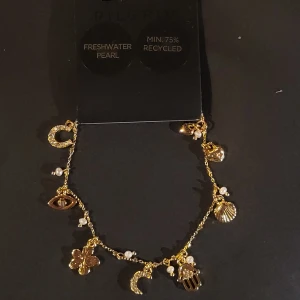 Gold plated  armband med berlocker från Pilgrim - OÖPPNAD Charmigt gold plated (rostar icke) armband från Pilgrim med flera berlocker som snäcka, blomma, öga, måne och hand. Armbandet är dekorerat med små sötvattenspärlor och har en tunn kedja. Perfekt accessoar för att lyfta din stil.