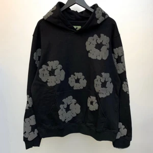 Svart denim tears hoodie  - Cool svart denim tears hoodie med stora gråa blommor tryckta över hela tröjan. Tröjan har huva och en klassisk magficka framtill. Perfekt för dig som vill sticka ut lite extra med ett unikt mönster.