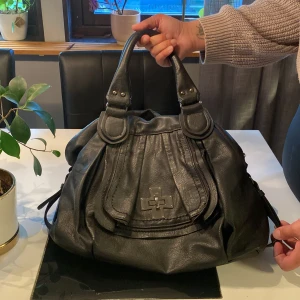Svart handväska med axelrem - Säljer en svart handväska i skinnimitation med både handtag och avtagbar axelrem. Väskan har en dragkedja på baksidan och ett stort fack med mönstrat innerfoder. Snygg detalj framtill och gott om plats för dina grejer.
