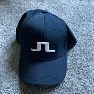 Navy keps från J.Lindeberg - Säljer en svart keps från J.Lindeberg med vit broderad logga framtill. Kepsen har böjd skärm och justerbar rem baktill för perfekt passform. Stilren och sportig design som passar till många tillfällen.