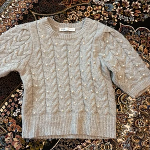 Grå stickad tröja med pärlor från Zara - Superfin grå stickad tröja från Zara med korta puffärmar och dekorativa pärlor framtill. Tröjan har ett klassiskt kabelstickat mönster och ribbade kanter. Perfekt för att lyfta din outfit med en söt detalj.