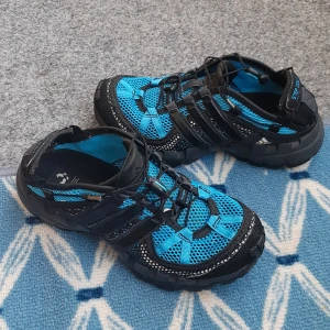 Blå och svarta sneakers från Adidas - Säljer ett par sportiga sneakers från Adidas i blått och svart med meshdetaljer och snörning. Skorna har robust sula och ventilerande material, perfekta för träning eller vardagsbruk. Märket syns tydligt på sidan och hälen. Skor är nästan nya,används några gånger. 