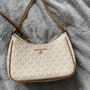 Michael Kors väska - Säljer en snygg beige axelväska från Michael Kors med guldiga kedjedetaljer och bruna kanter. Väskan har MK-logga över hela ytan, dragkedja upptill och ett innerfack. Perfekt storlek för det viktigaste och stilren design.