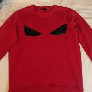 Röd sweatshirt från Fendi med svarta detaljer - Cool röd sweatshirt från Fendi med unika svarta ögon-detaljer framtill. Tröjan har rund hals och långa ärmar, perfekt för dig som vill sticka ut med en statement-piece. Mjuk bomullskänsla och snygg passform.