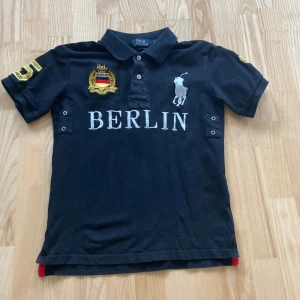 Svart pikétröja från Polo Ralph Lauren - Svart pikétröja från Polo Ralph Lauren med BERLIN-tryck och broderad logga framtill. Gula detaljer med siffran 5 på ryggen och ärmen. Klassisk krage och korta ärmar. Perfekt för en sportig och stilren look.