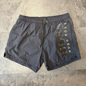 Svarta badshorts från Hugo Boss - Svarta badshorts från Hugo Boss med gulddetaljer och logga på vänster ben. Shortsen har snörning i midjan, innerbyxa i mesh och en bakficka med knapp och en lite innerficka. Perfekta för stranden eller poolen. 