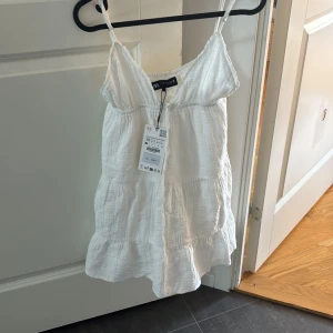 Vit kort klänning med smala axelband från ZARA - Säljer en söt vit kort klänning från ZARA i storlek XS. Klänningen har smala axelband, v-ringning och volangdetaljer nertill. Perfekt för varma dagar och till skolavslutningen.Ny med prislapp 