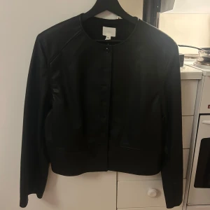 Svart skinnjacka från H&M - Säljer en svart skinnjacka från H&M i klassisk modell med rund halsringning och knäppning framtill. Jackan har lång ärm och en stilren, minimalistisk design som passar till många olika outfits. Storlek M, aldrig använd! Prislapp kvar 