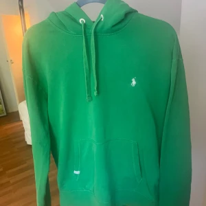 Grön hoodie från Polo Ralph Lauren - Säljer en grön hoodie från Polo Ralph Lauren med klassisk huva och dragsko. Tröjan har en broderad vit logga på bröstet och en stor magficka framtill. Perfekt för en avslappnad stil.